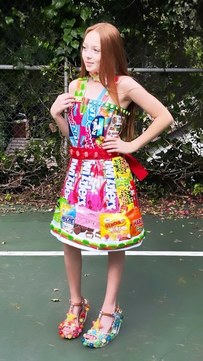 8-year-old fashion prodigy wears a creative dress made from colorful candy wrappers outdoors.