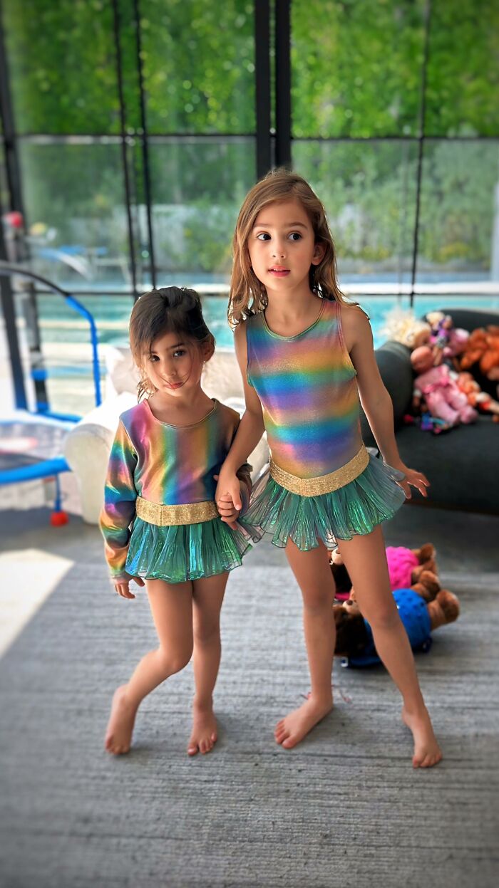 Two children in rainbow outfits designed by an 8-year-old fashion prodigy, standing indoors with toys in the background.