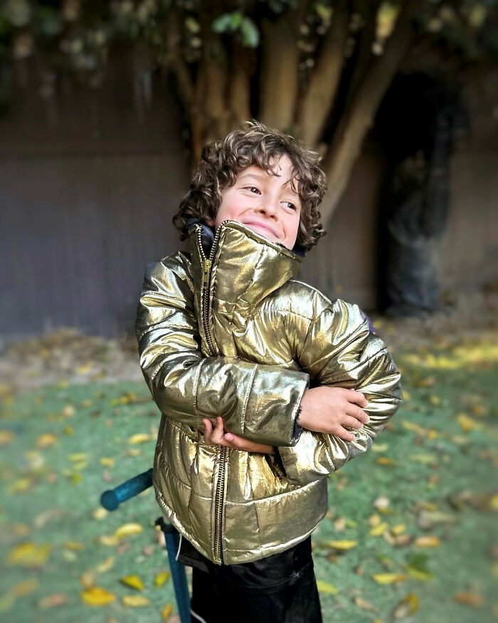 Young fashion prodigy in a shiny gold jacket, smiling outdoors amidst leafy surroundings.