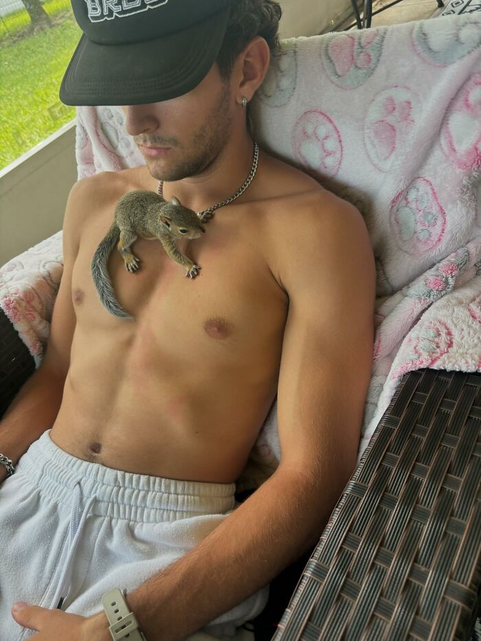 Man with a tiny squirrel resting on his chest, wearing a cap and relaxing on a patterned blanket.