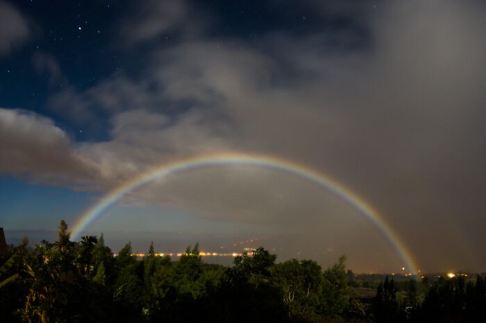 A moonbow, one of nature's incredible phenomena, arches over a night sky with scattered stars and glowing horizon.