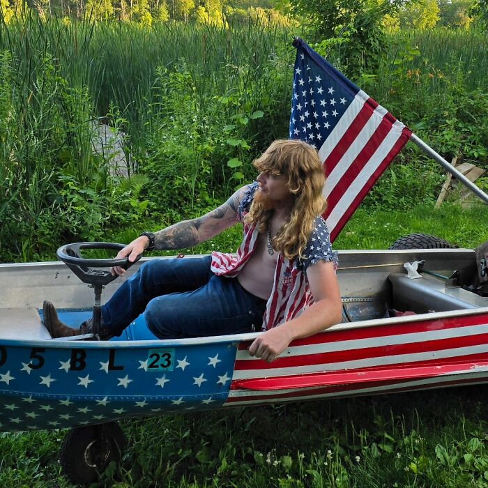 Participant with mullet hairstyle sits in a boat decorated with American flags, embracing bold hairdos.