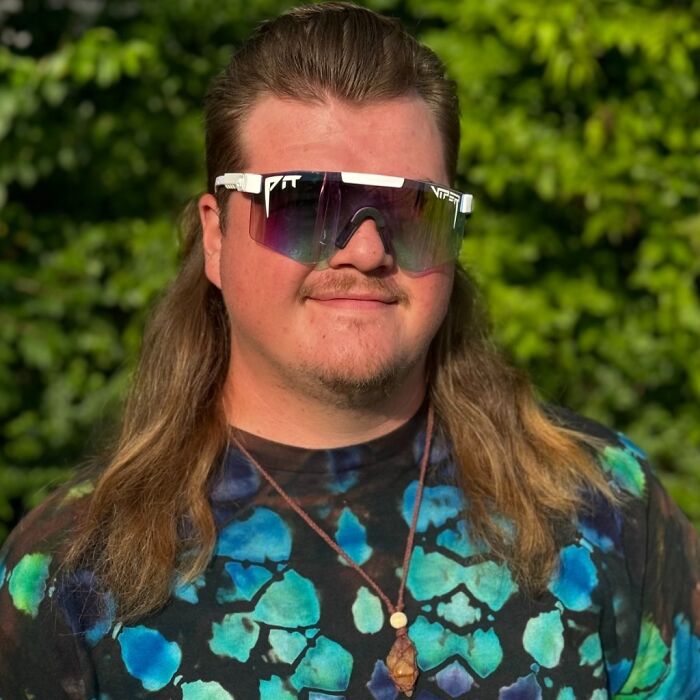 Man with mullet hairstyle and reflective sunglasses, wearing a colorful shirt, standing outdoors.