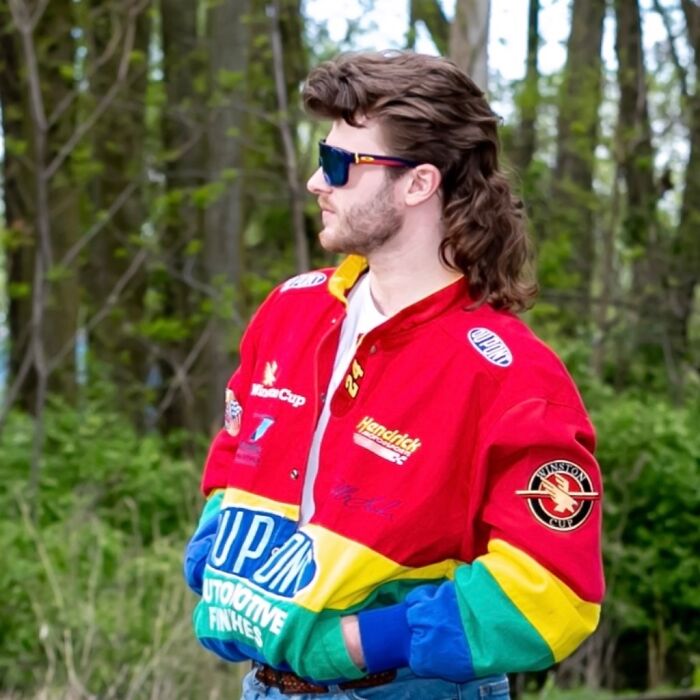 Man wearing sunglasses and a colorful jacket, showcasing a mullet hairstyle in a wooded area.