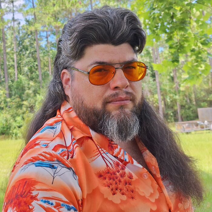 Man with a mullet hairstyle, wearing orange sunglasses and a floral shirt, standing outdoors.