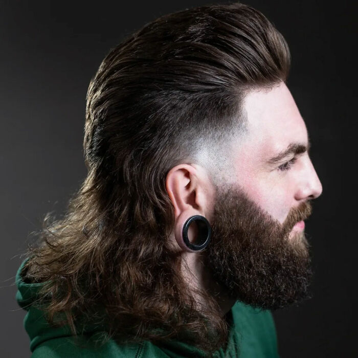 Man with stylish mullet hairstyle, featuring shaved sides and long back, wearing a green jacket.