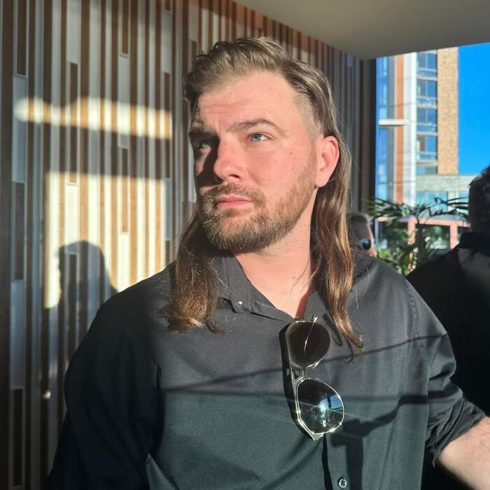 Man with mullet hairstyle standing indoors, wearing sunglasses on a black shirt.