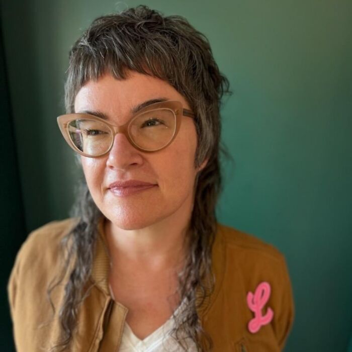 Person with glasses sporting a stylish mullet, wearing a brown jacket with a pink badge, green background.