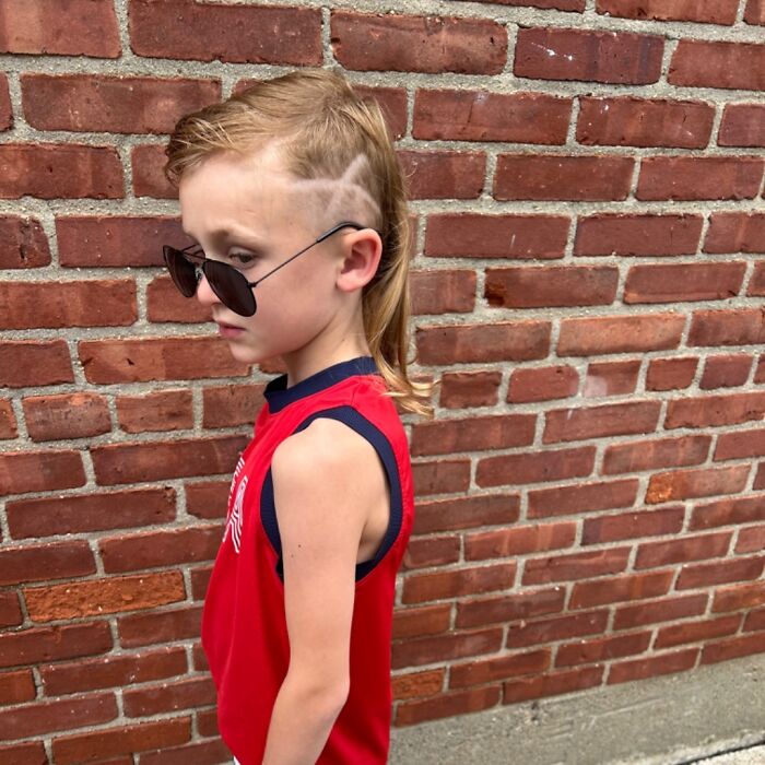 Child with a mullet hairstyle, wearing sunglasses and a red shirt, stands against a brick wall.