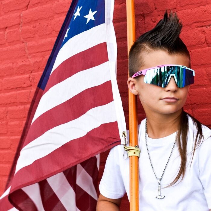 Young person with stylish mullet, wearing sunglasses, holding an American flag, standing against a red wall.