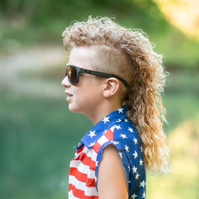 Kid with a mullet hairstyle, wearing sunglasses and an American flag shirt, outdoors.