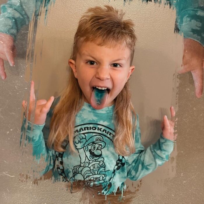 Child with a mullet hairstyle, wearing a Mario Kart shirt, sticking out a tongue dyed blue.