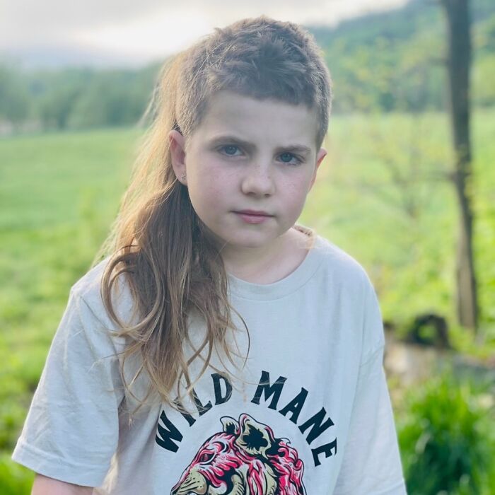 Child with a mullet hairstyle outdoors, wearing a "Wild Mane" shirt, showcasing a trendy 2024 hairdo.