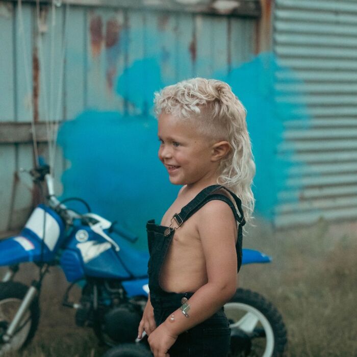 Child with a mullet hairstyle standing next to a blue dirt bike, smiling outdoors.