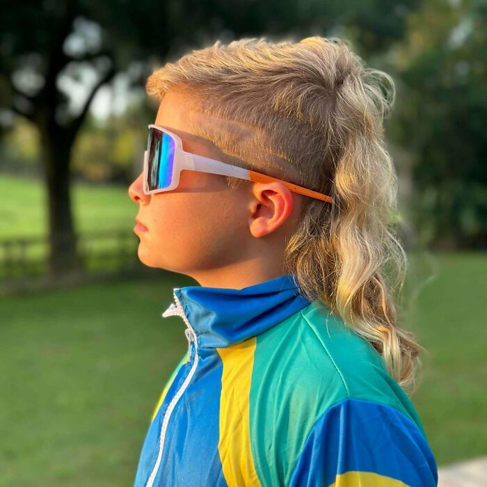 Child with mullet hairstyle wearing colorful jacket and large sunglasses, standing outdoors.