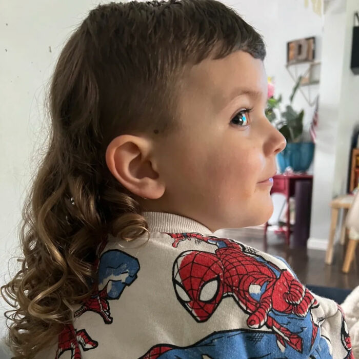 Young child with a mullet hairstyle, wearing a Spiderman shirt, looking sideways in a cozy room.