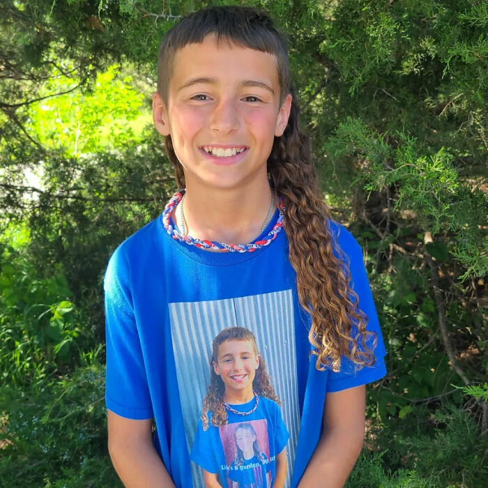 Child smiling with a mullet hairstyle, wearing a blue shirt featuring the same hairstyle, outdoors.