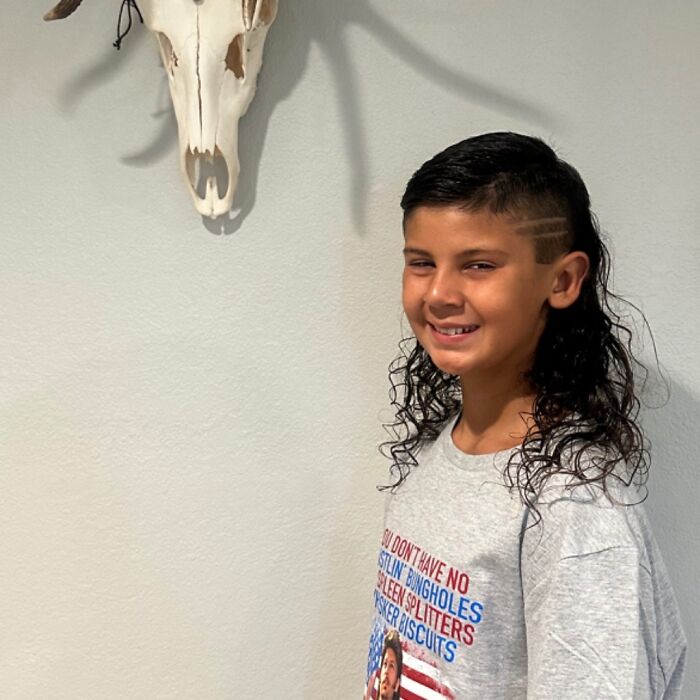 Kid with mullet hairstyle smiling beside a wall-mounted skull, wearing a graphic tee.