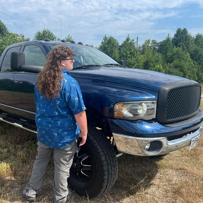 Person with mullet hairstyle standing near a blue truck in an outdoor setting.