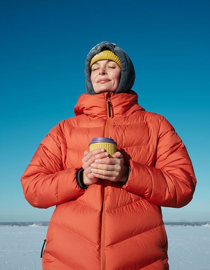 Person in orange coat enjoying a sunny winter day, holding a cup, illustrating Norwegian secret to staying warm.