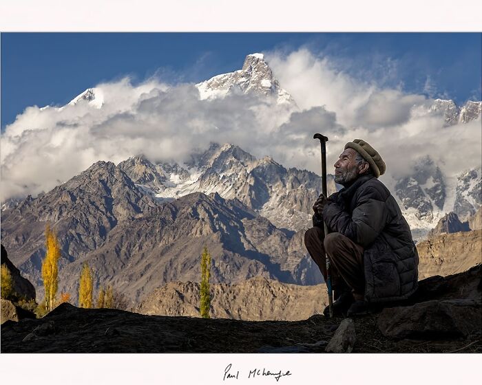 Man with a walking stick gazing at mountains under a cloudy sky, captured by photographer Paul McKenzie.