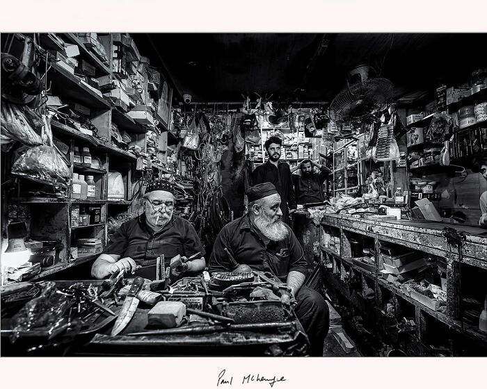 Inside a cluttered workshop, two men sit surrounded by tools, photographed by Paul McKenzie.