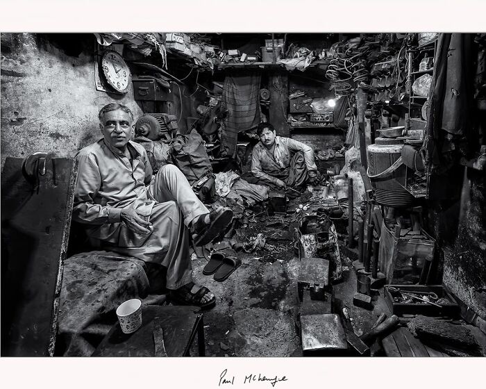 Two men in a cluttered workshop, captured in black and white by Paul McKenzie.