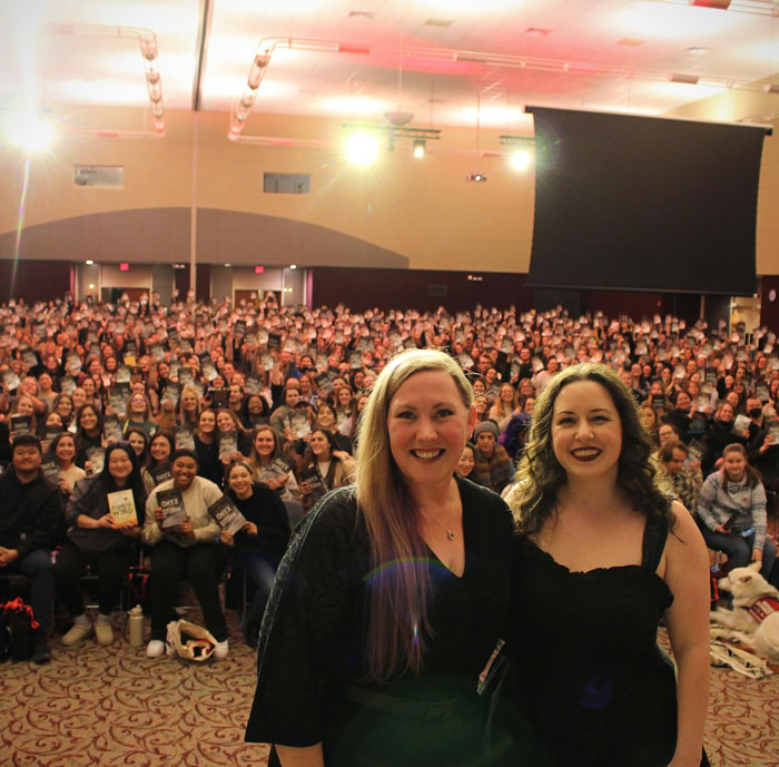 Two authors smiling in front of a large audience holding spicy romantasy novels at a book event.