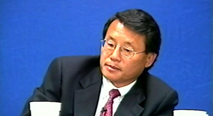 Man in a suit and glasses sitting against a blue background, appearing thoughtful.