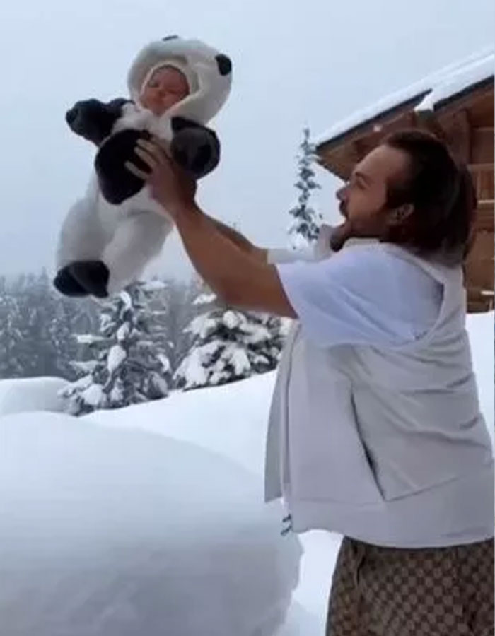 Influencer dad holding baby in panda suit amid snowy landscape, with snow-covered trees and cabin in the background.