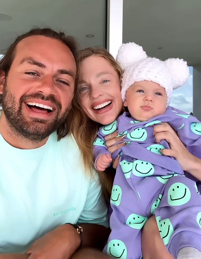 Family posing happily, dad in a mint shirt, mom holding baby in a purple outfit and white hat, showcasing influencer life.