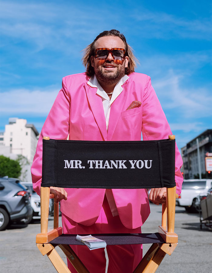 Man in a pink suit with sunglasses leaning on a director's chair that reads "Mr. Thank You," outdoors under a blue sky.