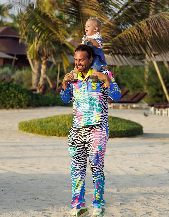 Influencer dad on a beach with their child on shoulders, dressed in colorful attire.