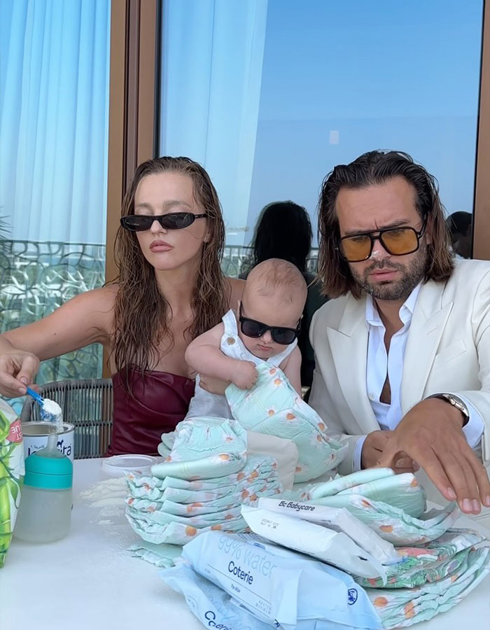 Influencer dad with baby, both in sunglasses, sitting at a table with diapers and baby supplies.