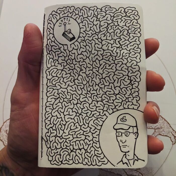 Hand holding a notebook with funny drawing of a maze and a man in a cap, by Ed Harrington.