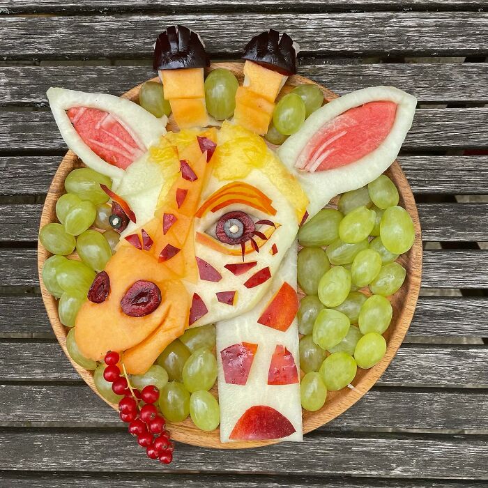 Fruit art of a giraffe made from melon, grapes, and berries on a wooden table, showcasing healthy eating creativity.