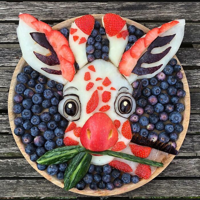 Healthy eating art made from fruit, featuring a creative animal face with blueberries, apples, and strawberries on a wooden plate.