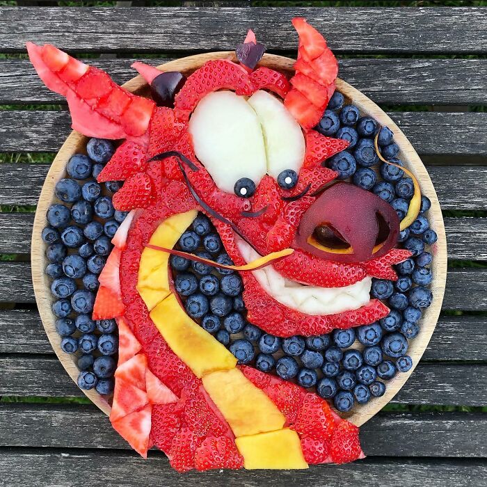 Healthy eating art featuring a dragon face made from strawberries, blueberries, and assorted fruits on a wooden surface.