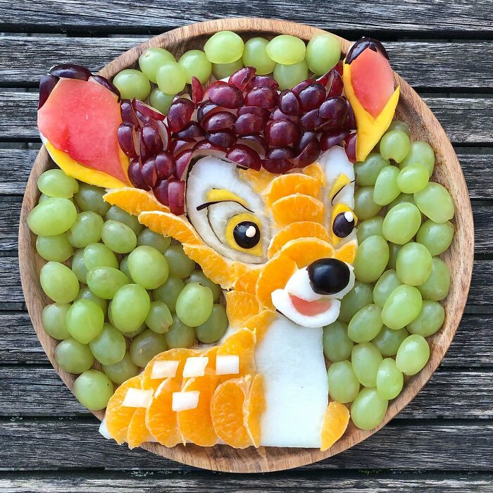 Healthy eating art featuring a fruit arrangement resembling a cartoon character with grapes, oranges, and apples on a wooden plate.