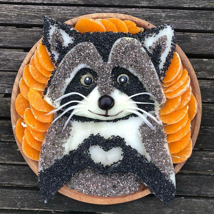 Raccoon art made from healthy foods like fruit and seeds on a wooden plate.