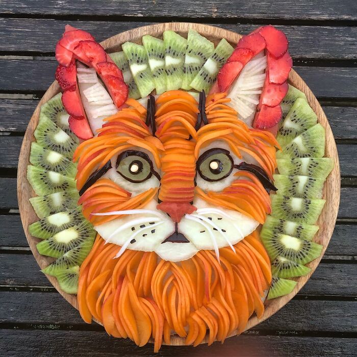 Healthy eating art featuring a cat face made from sliced fruits and vegetables on a wooden platter.