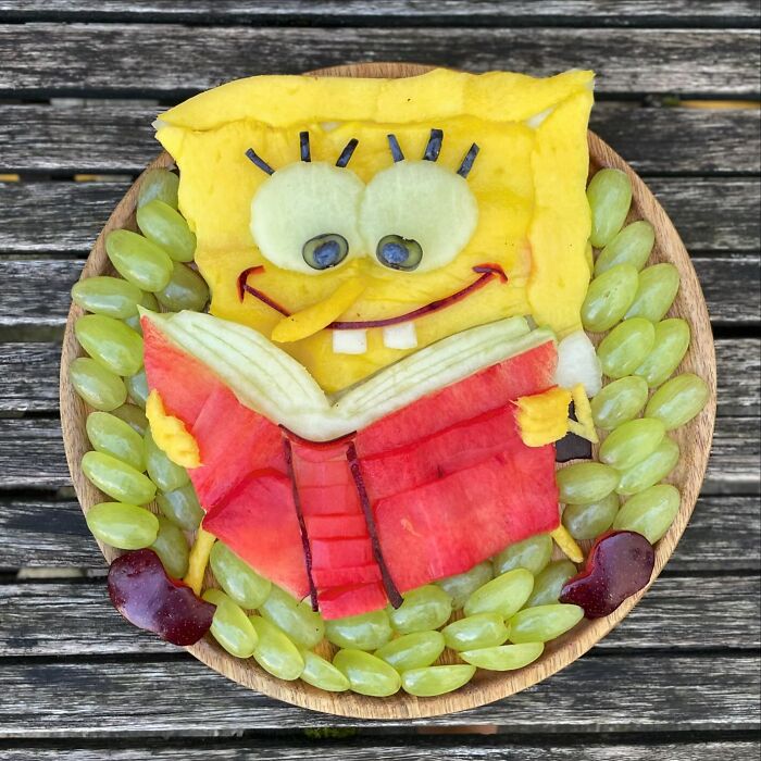 Fruit art depicting a cartoon character made from melon, grapes, and berries on a wooden table.