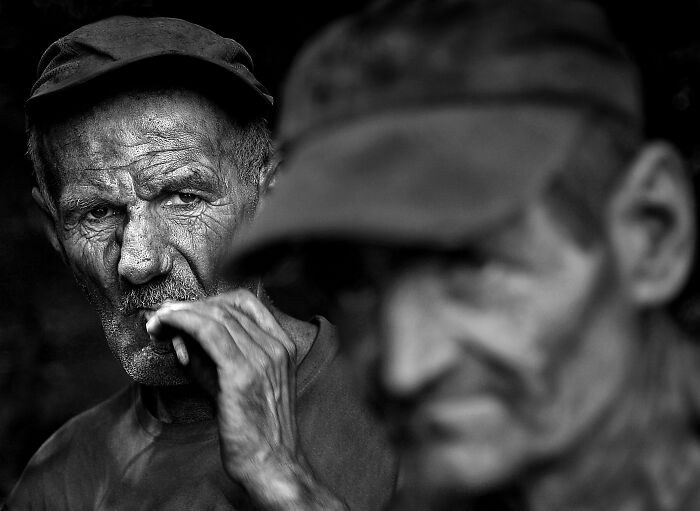 Two men in hats, one in focus with intense gaze, capturing the resilience and beauty of everyday people in black and white.