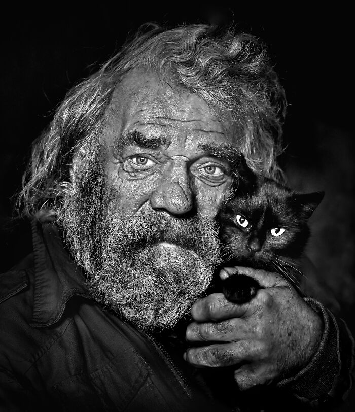 A man with a weathered face holds a black cat, showcasing resilience and beauty in everyday life.