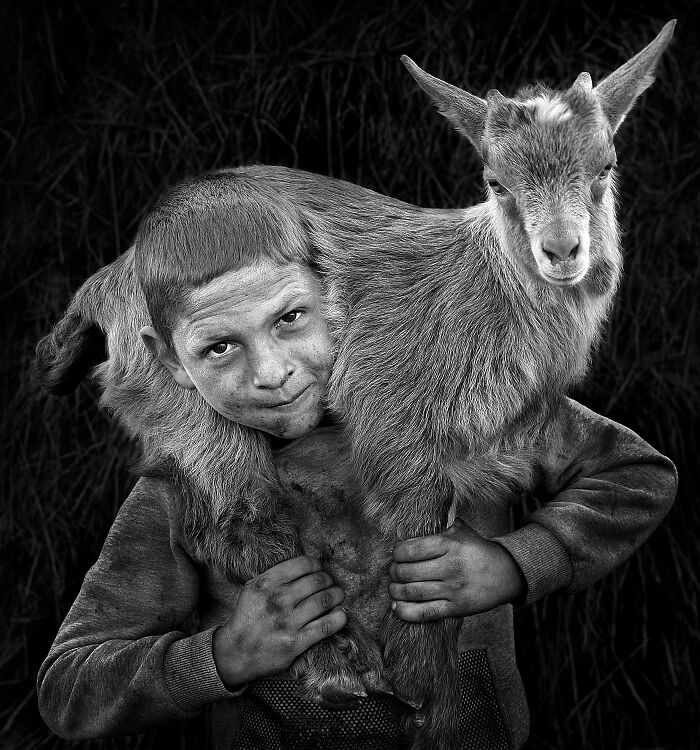 A young boy smiling, carrying a goat on his shoulders, showcasing resilience and beauty of everyday people.