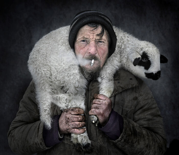 A rugged man with a lamb draped over his shoulders, showcasing resilience and beauty captured by Istvan Kerekes.