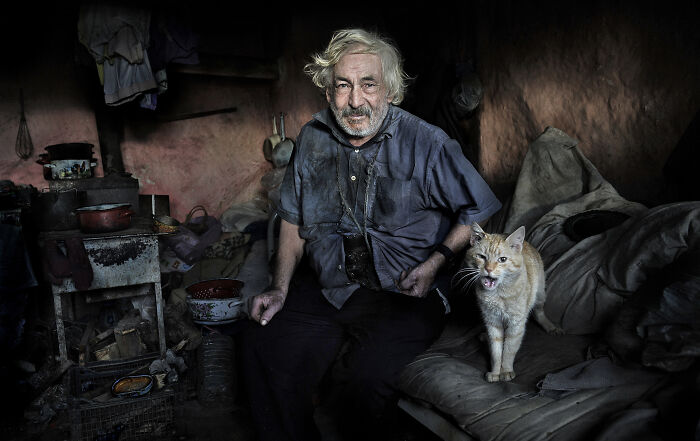 An elderly man with a cat in a rustic room, illustrating resilience and beauty of everyday people.