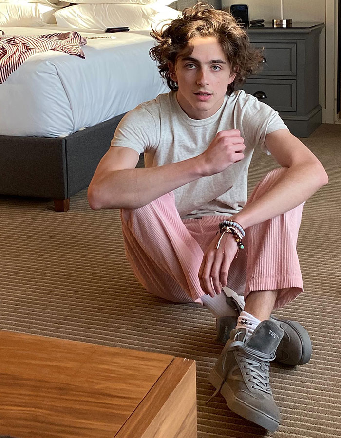 Young man seated indoors, wearing a casual outfit, includes pink pants and grey sneakers.