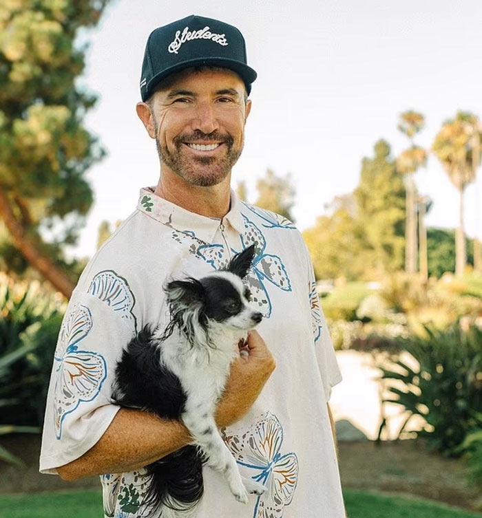 Man in a floral shirt and cap holding a small dog in a sunny garden setting, related to MySpace founder.