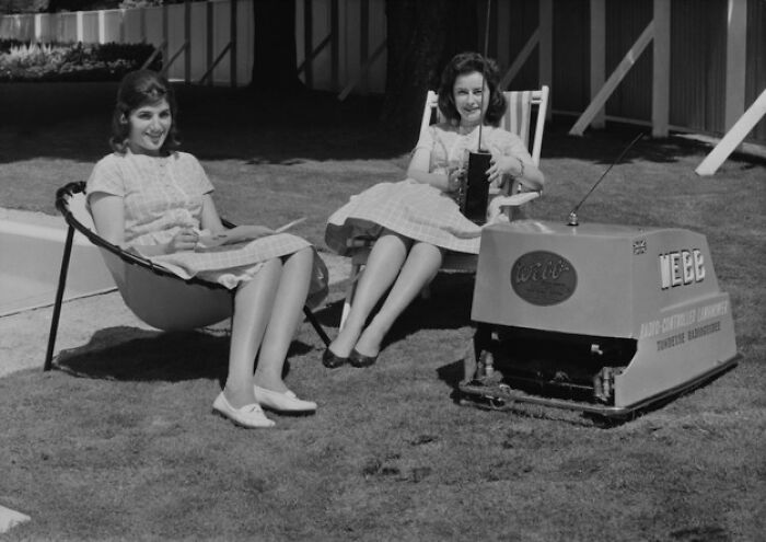 Two women sitting on chairs, outdoors, beside an early automated lawnmower, a historical invention ahead of its time.